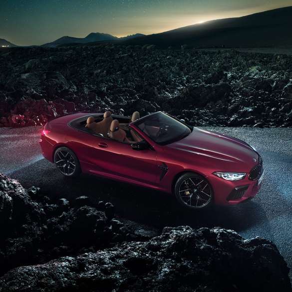 BMW Μ8 Competition Cabrio, πλαϊνή λήψη τριών τετάρτων. BMW Μ8 Competition Cabrio σε κόκκινο Motegi μεταλλικό με ανοικτή υφασμάτινη οροφή, πλαϊνή λήψη τριών τετάρτων, σταθμευμένη στην άσφαλτο τη νύχτα.