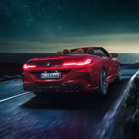 BMW Μ8 Competition Cabrio, πίσω λήψη τριών τετάρτων. BMW Μ8 Competition Cabrio σε κόκκινο Motegi μεταλλικό με ανοικτή υφασμάτινη οροφή, πίσω λήψη τριών τετάρτων, κινούμενη σε παράκτιο δρόμο τη νύχτα.