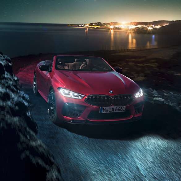 BMW Μ8 Competition Cabrio, μπροστινή λήψη τριών τετάρτων. BMW Μ8 Competition Cabrio σε κόκκινο Motegi μεταλλικό με ανοικτή υφασμάτινη οροφή, μπροστινή λήψη τριών τετάρτων, κινούμενη σε παράκτιο δρόμο τη νύχτα.