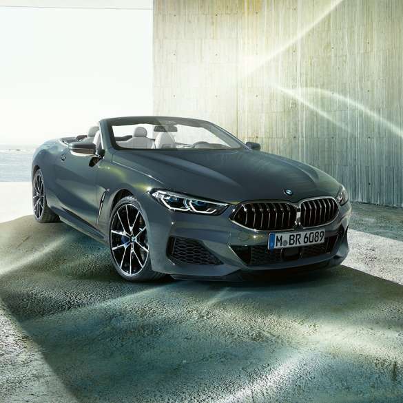 BMW M850i xDrive Cabrio, μπροστινή λήψη τριών τετάρτων. BMW M850i xDrive Cabrio σε γκρι Dravit μεταλλικό με ανοικτή υφασμάτινη οροφή, μπροστινή λήψη τριών τετάρτων, σταθμευμένη μπροστά από ακτή.