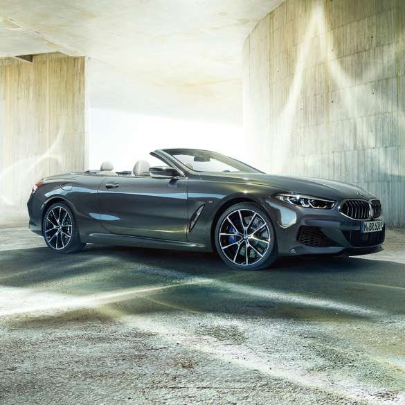 BMW M850i xDrive Cabrio, πλαϊνή λήψη τριών τετάρτων. BMW M850i xDrive Cabrio σε γκρι Dravit μεταλλικό με ανοικτή υφασμάτινη οροφή, πλαϊνή λήψη τριών τετάρτων, σταθμευμένη μπροστά από τσιμεντένιο κτίριο.