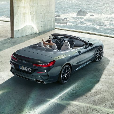BMW M850i xDrive Cabrio, πανοραμική λήψη. BMW M850i xDrive Cabrio σε γκρι Dravit μεταλλικό με ανοικτή υφασμάτινη οροφή, πανοραμική λήψη, σταθμευμένη σε τσιμεντένιο κτίριο, μπροστά από ωκεανό.