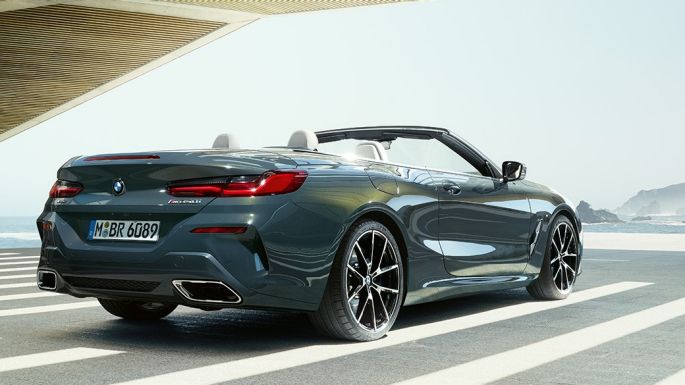 BMW M850i xDrive Cabrio, πίσω λήψη τριών τετάρτων. BMW M850i xDrive Cabrio σε γκρι Dravit μεταλλικό με ανοικτή υφασμάτινη οροφή, πίσω λήψη τριών τετάρτων, σταθμευμένη μπροστά σε ωκεανό.