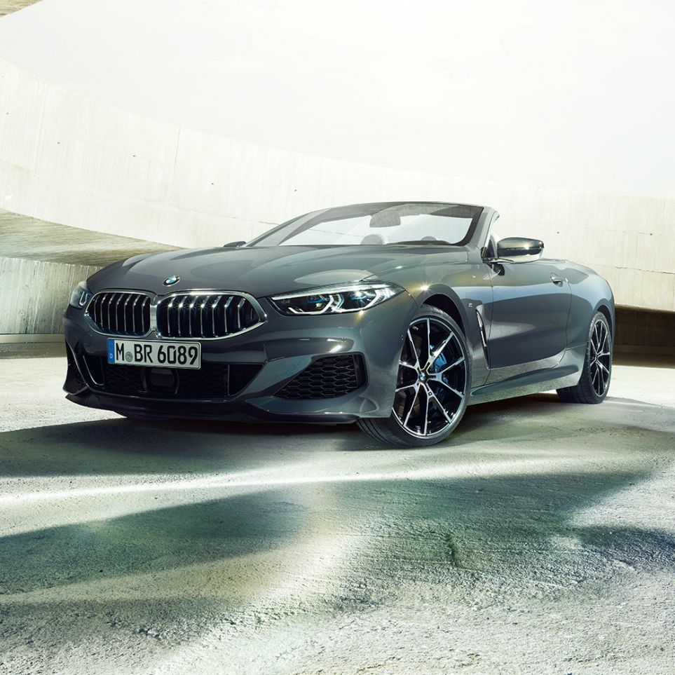 BMW M850i xDrive Cabrio, μπροστινή λήψη τριών τετάρτων. BMW M850i xDrive Cabrio σε γκρι Dravit μεταλλικό με ανοικτή υφασμάτινη οροφή, μπροστινή λήψη τριών τετάρτων, σταθμευμένη μπροστά από τσιμεντένιο κτίριο.