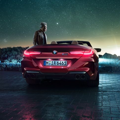 BMW M8 Competition Cabrio, πίσω λήψη. BMW Μ8 Competition Cabrio σε κόκκινο Motegi μεταλλικό με ανοικτή υφασμάτινη οροφή, πίσω λήψη, σταθμευμένη, με άντρα κάτω από έναστρο ουρανό.
