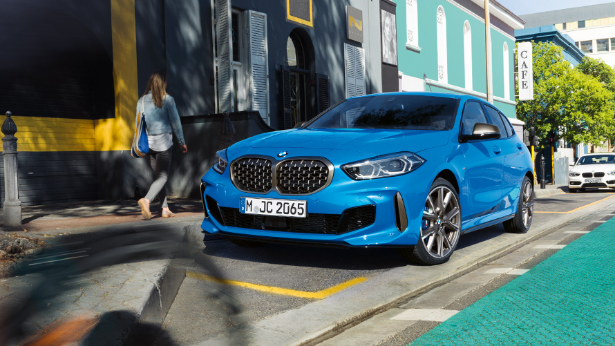 BMW Σειρά 1, F40, σε μπλε Misano μεταλλικό, μπροστινή λήψη τριών τετάρτων BMW Σειρά 1, F40 σε μπλε Misano μεταλλικό, μπροστινή λήψη τριών τετάρτων, σταθμευμένη στην άκρη του δρόμου με γυναίκα