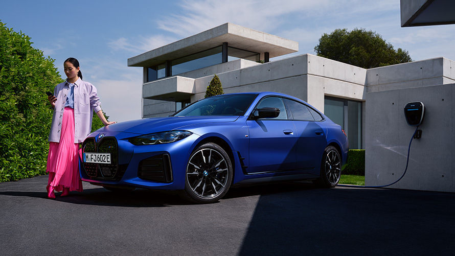 Πλαϊνή λήψη τριών τετάρτων, BMW i4 Μ50, G26, 2021, με γυναίκα μπροστά από σπίτι BMW i4 M50, G26, 2021, BMW Individual μπλε Frozen Portimao μεταλλικό, πλαϊνή λήψη τριών τετάρτων με γυναίκα μπροστά από σπίτι