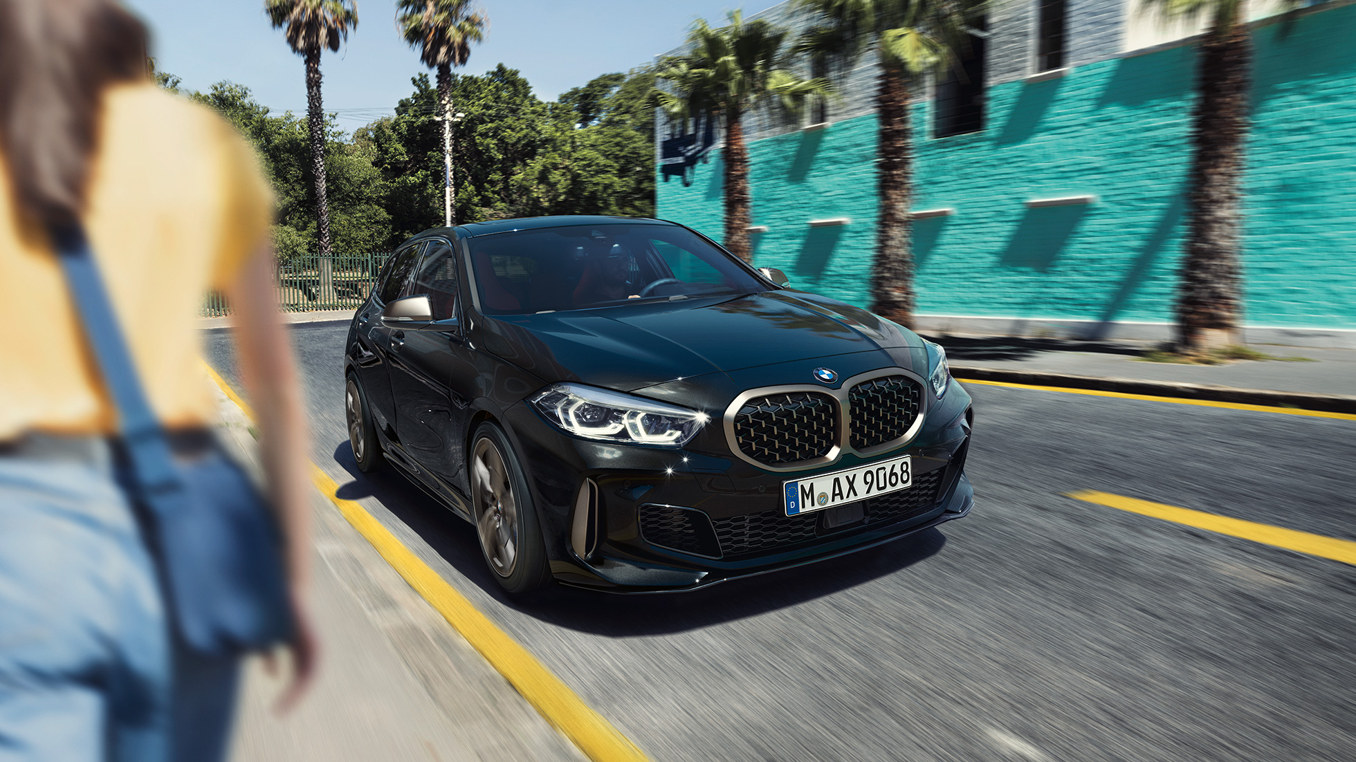 BMW M135i xDrive, μπροστινή λήψη τριών τετάρτων. BMW M135i xDrive σε μαύρο Sapphire μεταλλικό, μπροστινή λήψη τριών τετάρτων, σε κίνηση, με γυναίκα στο προσκήνιο.