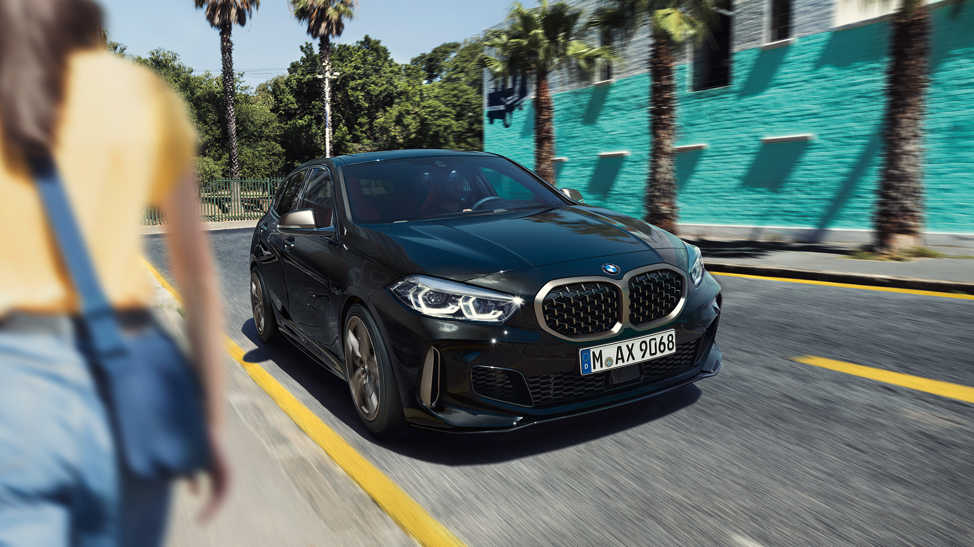 BMW M135i xDrive, μπροστινή λήψη τριών τετάρτων. BMW M135i xDrive σε μαύρο Sapphire μεταλλικό, μπροστινή λήψη τριών τετάρτων, σε κίνηση, με γυναίκα στο προσκήνιο.
