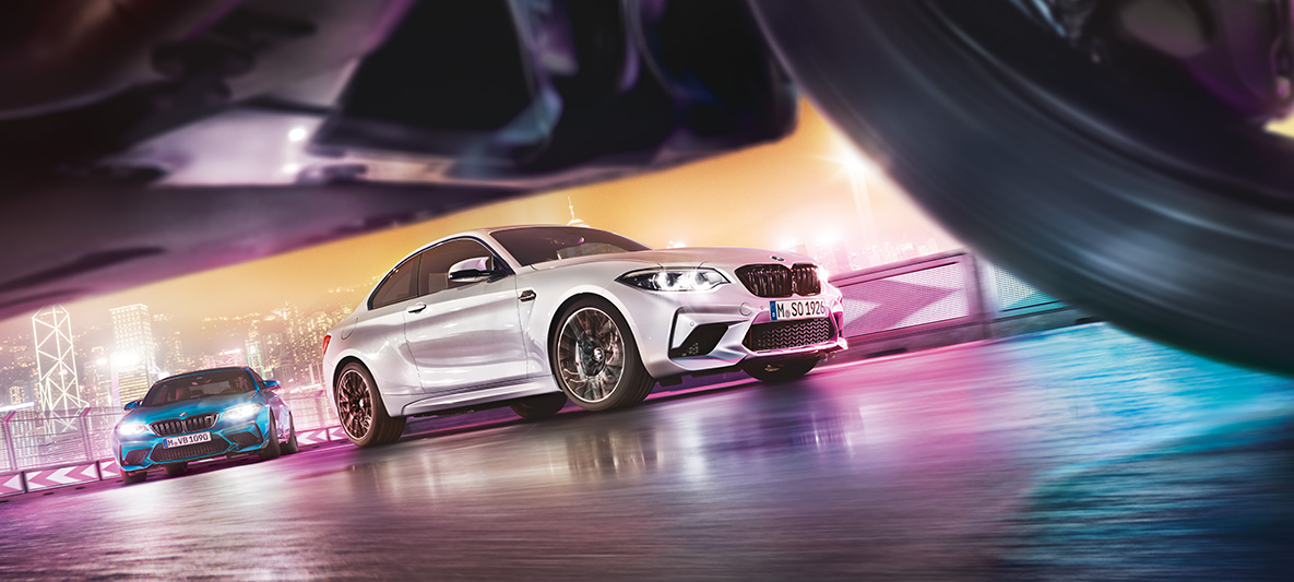 Η BMW M2 Competition σε άσφαλτο με αντανακλάσεις. Δύο BMW M2 Competition στην αγωνιστική πίστα μπροστά από νυχτερινό αστικό τοπίο, λήψη από το οδόστρωμα.