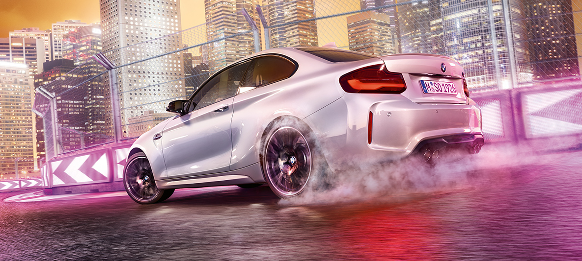 Η BMW M2 Competition επιταχύνει Μία λευκή BMW M2 Competition ντριφτάρει μέσα στην αγωνιστική πίστα, μπροστά από τον νυχτερινό αστικό ορίζοντα.