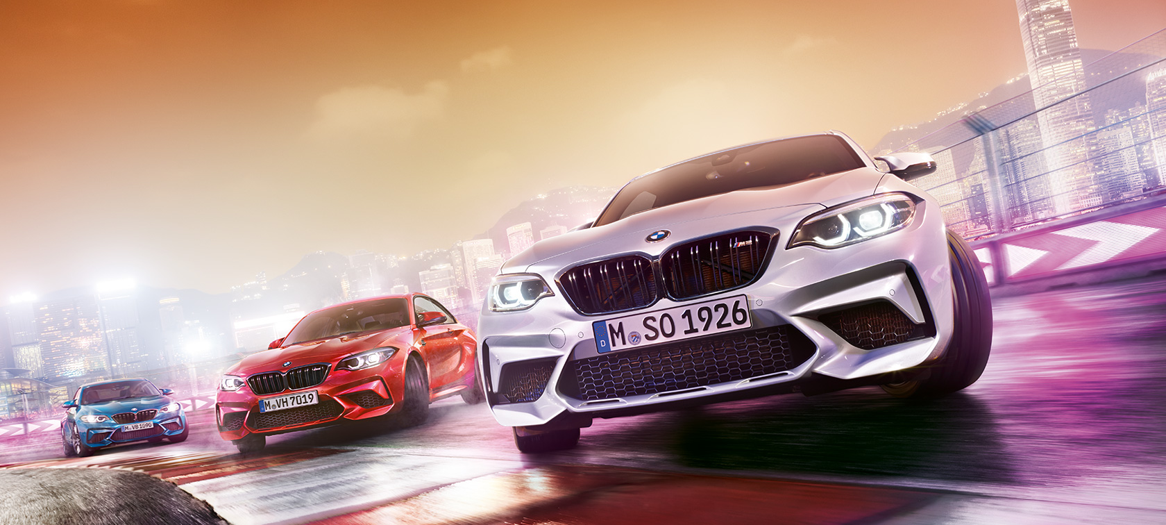 BMW M2 Competition σε δράση Τρεις BMW M2 Competition ντριφτάρουν μέσα στην αγωνιστική πίστα με φόντο το νυχτερινό αστικό τοπίο.