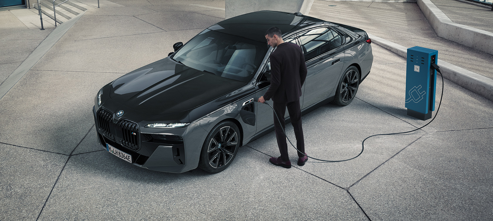 BMW M760e xDrive Sedan, G70, πακέτο M Performance BMW M760e xDrive Sedan, G70, πακέτο M Performance, χρώμα αμαξώματος BMW Individual μαύρο Sapphire σε διχρωμία, BMW Individual γκρι Dravite μεταλλικό, μπροστινή λήψη τριών τετάρτων, άντρας με καλώδιο φόρτισης δίπλα από σταθμό φόρτισης