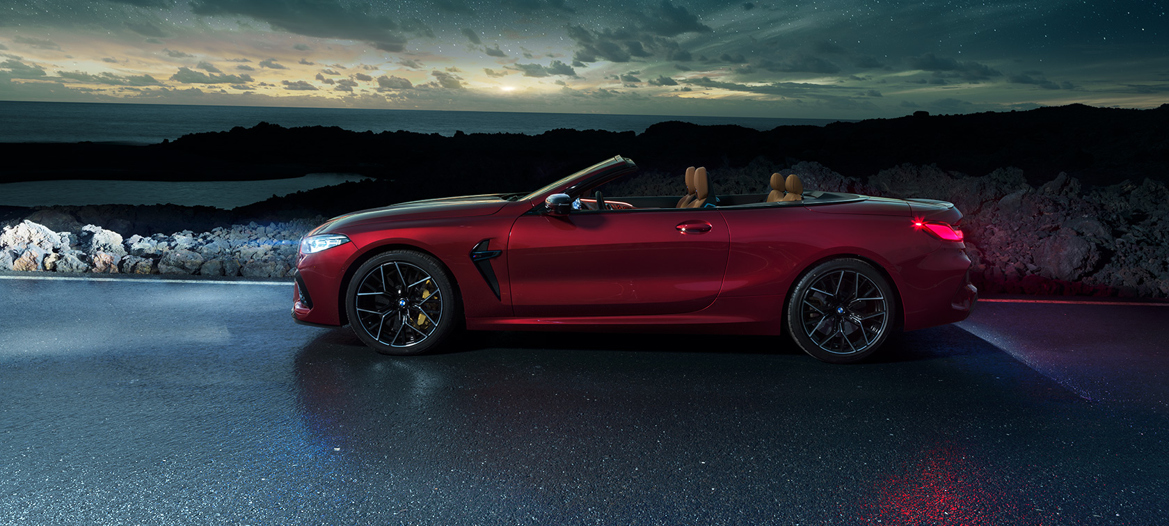 BMW M8 Competition Cabrio, πλαϊνή λήψη. BMW Μ8 Competition Cabrio σε κόκκινο Motegi μεταλλικό με ανοικτή υφασμάτινη οροφή, πλαϊνή λήψη, σταθμευμένη σε παράκτιο δρόμο κάτω από έναστρο ουρανό.