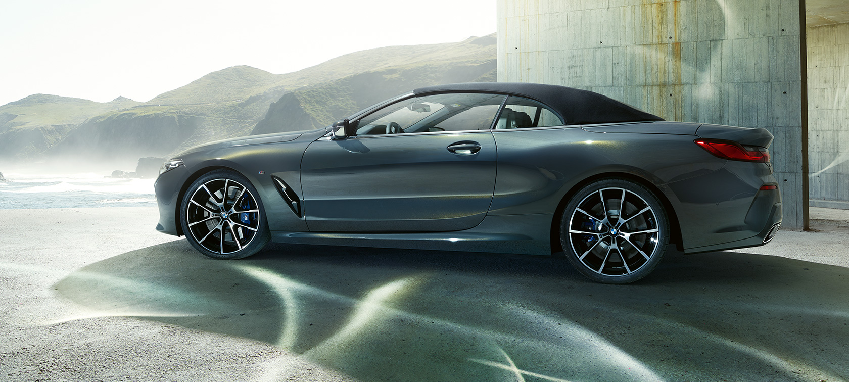 BMW M850i xDrive Cabrio, πλαϊνή λήψη. BMW M850i xDrive Cabrio σε γκρι Dravit μεταλλικό με κλειστή υφασμάτινη οροφή, πλαϊνή λήψη μπροστά από λόφους.