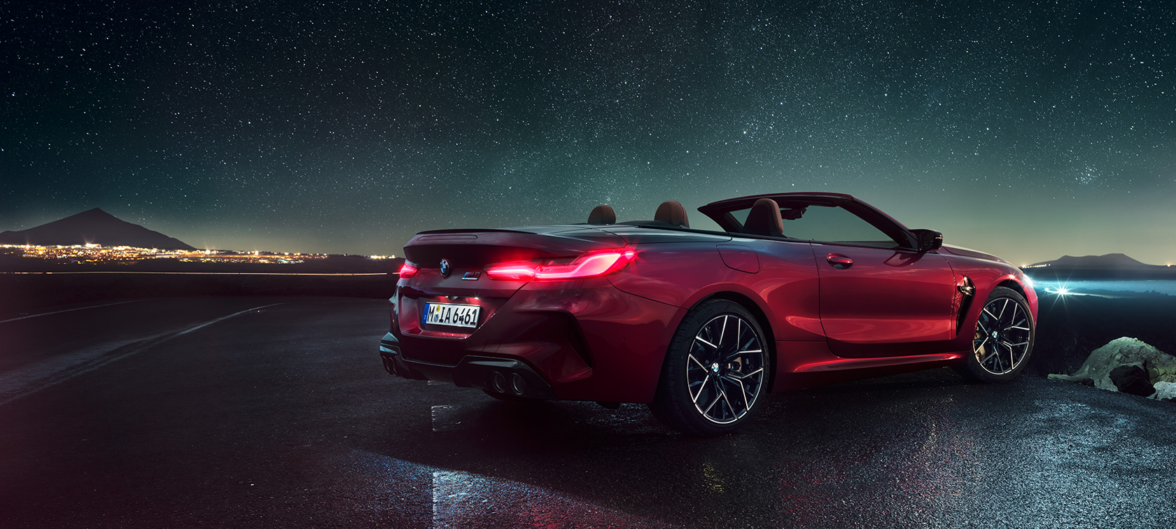 BMW Μ8 Competition Cabrio, πίσω λήψη τριών τετάρτων. BMW Μ8 Competition Cabrio σε κόκκινο Motegi μεταλλικό με ανοικτή υφασμάτινη οροφή, πίσω λήψη τριών τετάρτων, σταθμευμένη κάτω από έναστρο ουρανό.