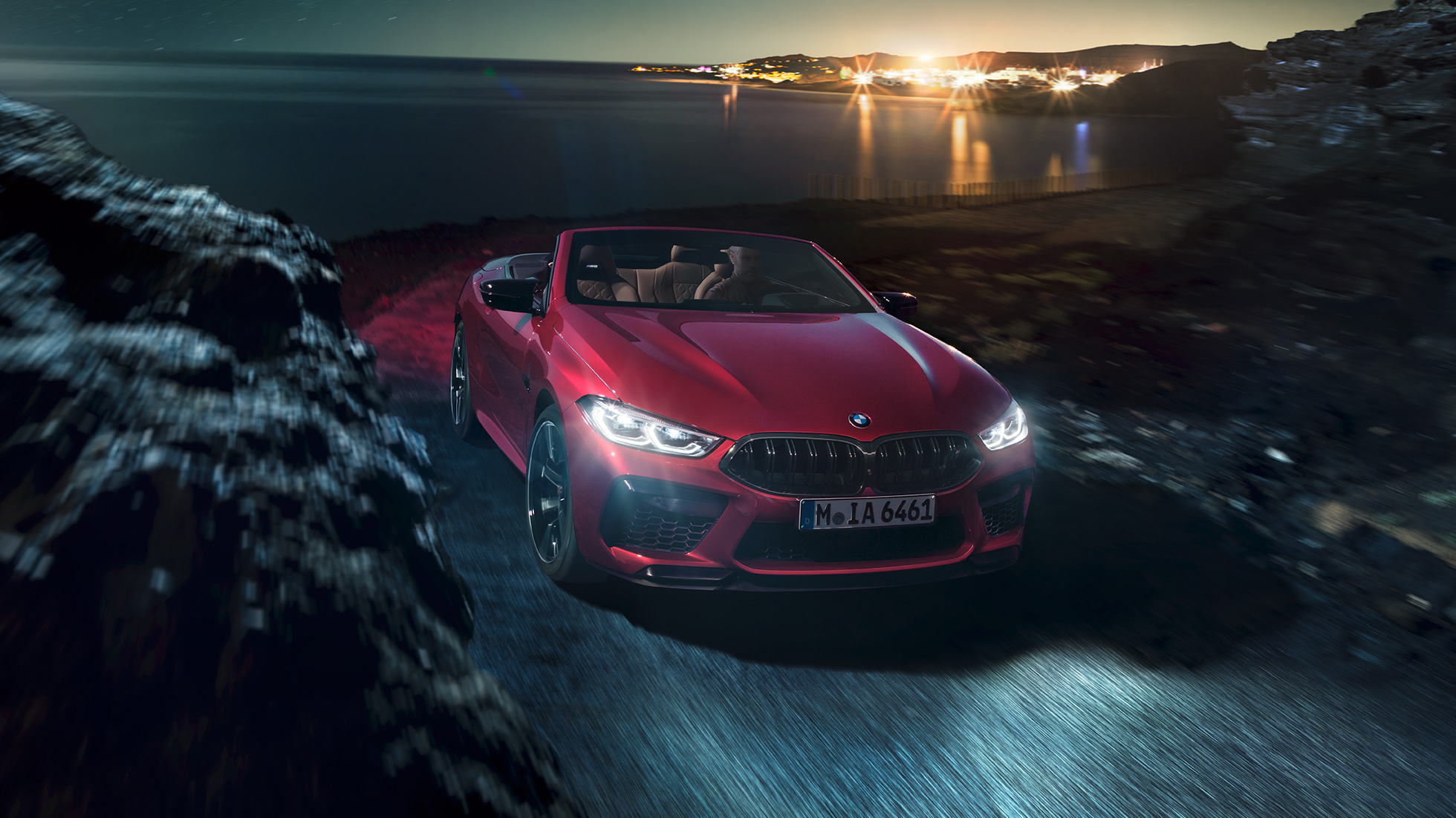 BMW Μ8 Competition Cabrio, μπροστινή λήψη τριών τετάρτων. BMW Μ8 Competition Cabrio σε κόκκινο Motegi μεταλλικό με ανοικτή υφασμάτινη οροφή, μπροστινή λήψη τριών τετάρτων, κινούμενη σε παράκτιο δρόμο τη νύχτα.