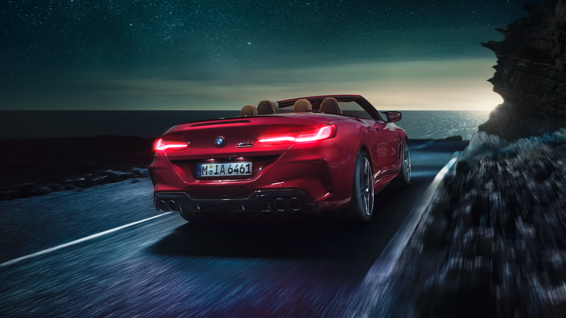 BMW Μ8 Competition Cabrio, πίσω λήψη τριών τετάρτων. BMW Μ8 Competition Cabrio σε κόκκινο Motegi μεταλλικό με ανοικτή υφασμάτινη οροφή, πίσω λήψη τριών τετάρτων, κινούμενη σε παράκτιο δρόμο τη νύχτα.
