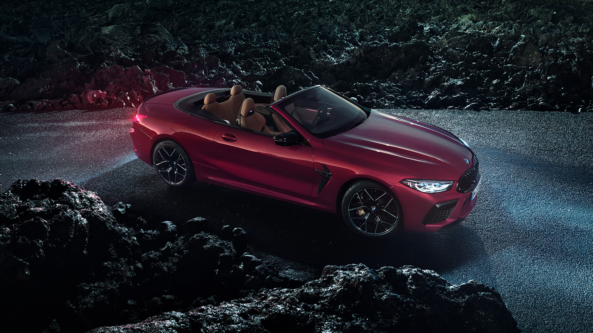 BMW Μ8 Competition Cabrio, πλαϊνή λήψη τριών τετάρτων. BMW Μ8 Competition Cabrio σε κόκκινο Motegi μεταλλικό με ανοικτή υφασμάτινη οροφή, πλαϊνή λήψη τριών τετάρτων, σταθμευμένη στην άσφαλτο τη νύχτα.