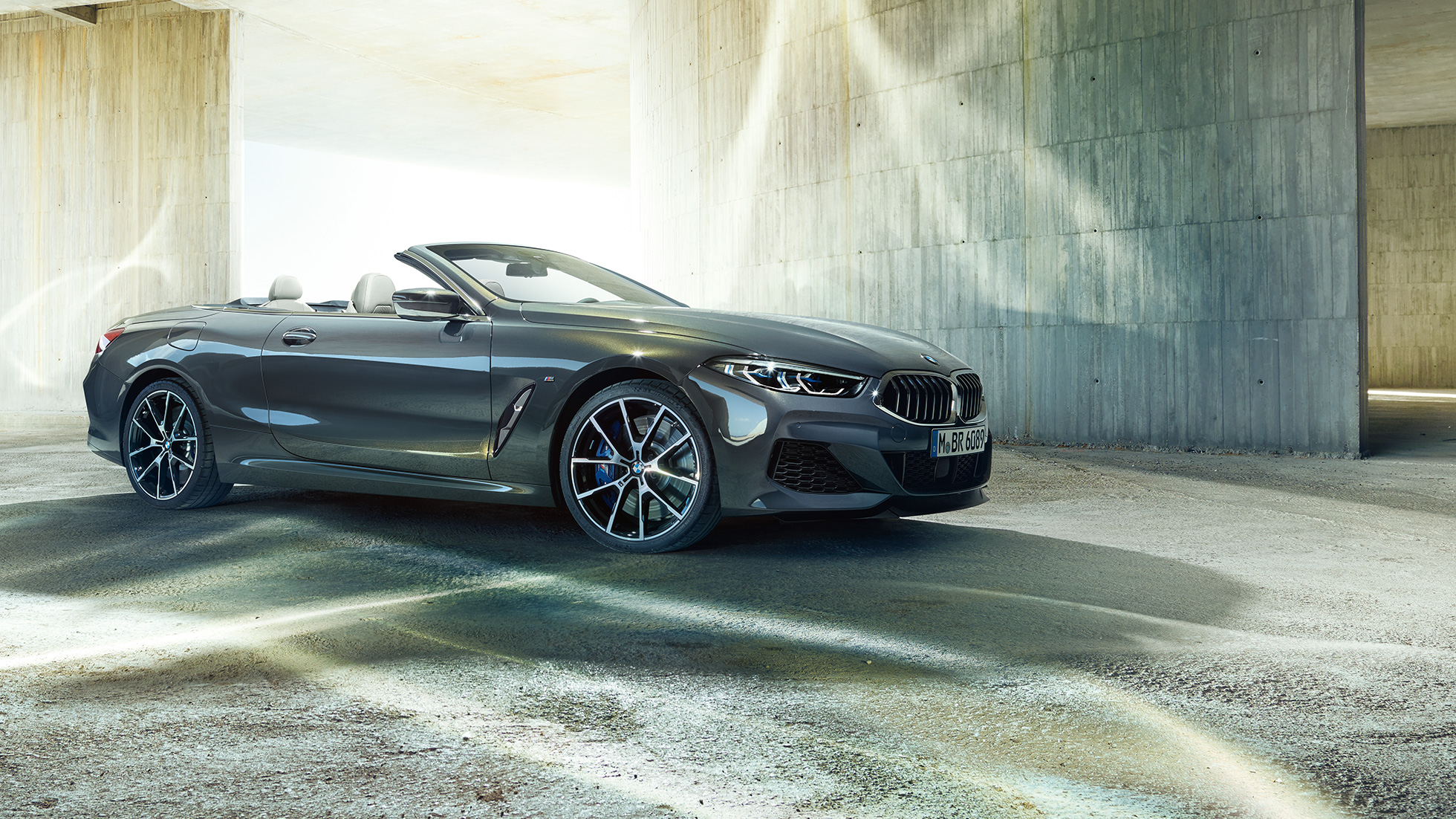 BMW M850i xDrive Cabrio, πλαϊνή λήψη τριών τετάρτων. BMW M850i xDrive Cabrio σε γκρι Dravit μεταλλικό με ανοικτή υφασμάτινη οροφή, πλαϊνή λήψη τριών τετάρτων, σταθμευμένη μπροστά από τσιμεντένιο κτίριο.