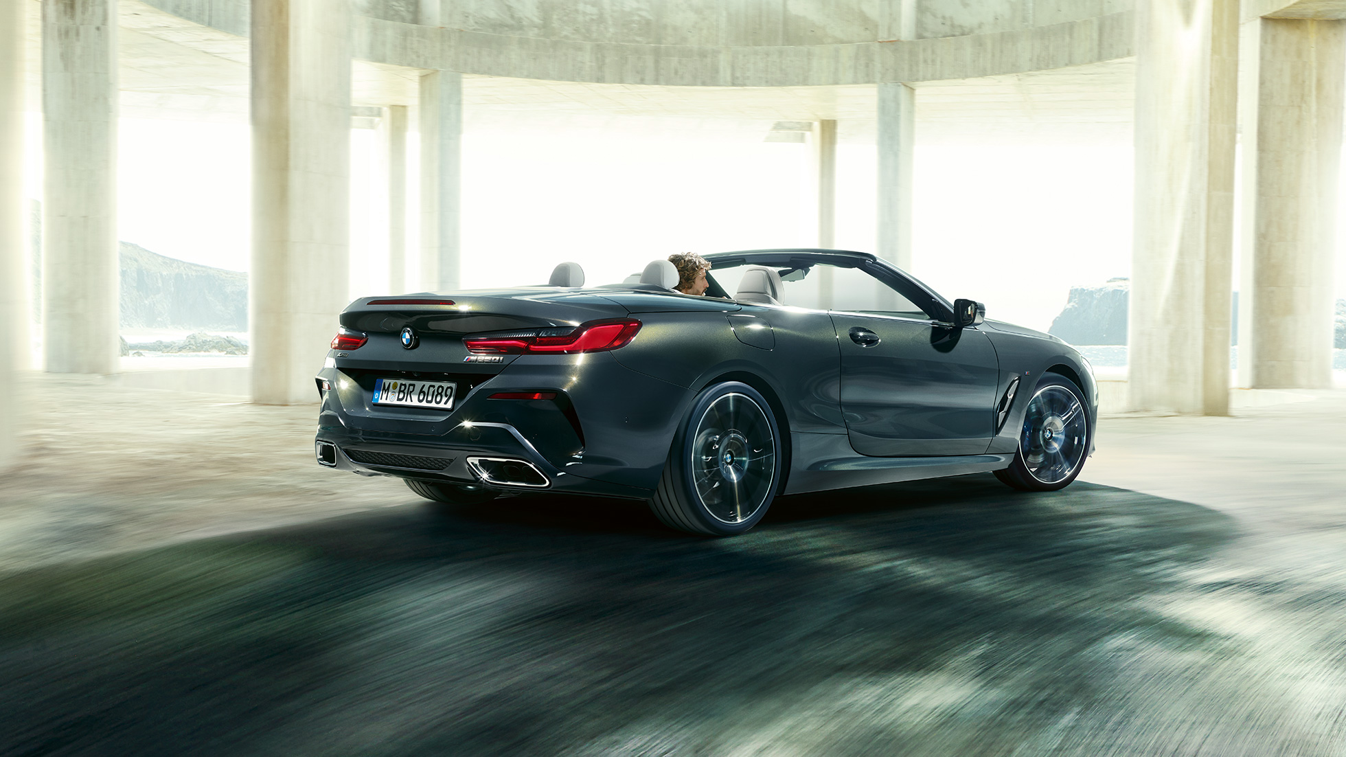 BMW M850i xDrive Cabrio, πίσω λήψη τριών τετάρτων. BMW M850i xDrive Cabrio σε γκρι Dravit μεταλλικό με ανοικτή υφασμάτινη οροφή, πίσω λήψη τριών τετάρτων, διερχόμενη μέσα από τσιμεντένιο κτίριο.