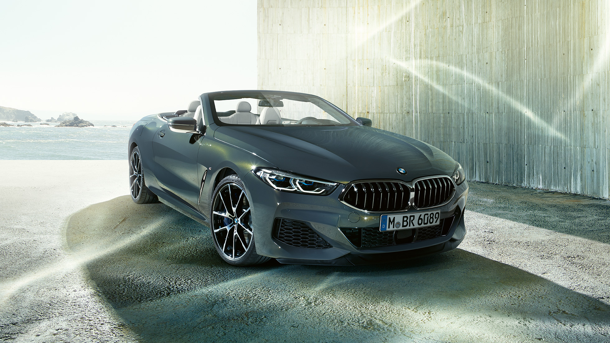 BMW M850i xDrive Cabrio, μπροστινή λήψη τριών τετάρτων. BMW M850i xDrive Cabrio σε γκρι Dravit μεταλλικό με ανοικτή υφασμάτινη οροφή, μπροστινή λήψη τριών τετάρτων, σταθμευμένη μπροστά από ακτή.