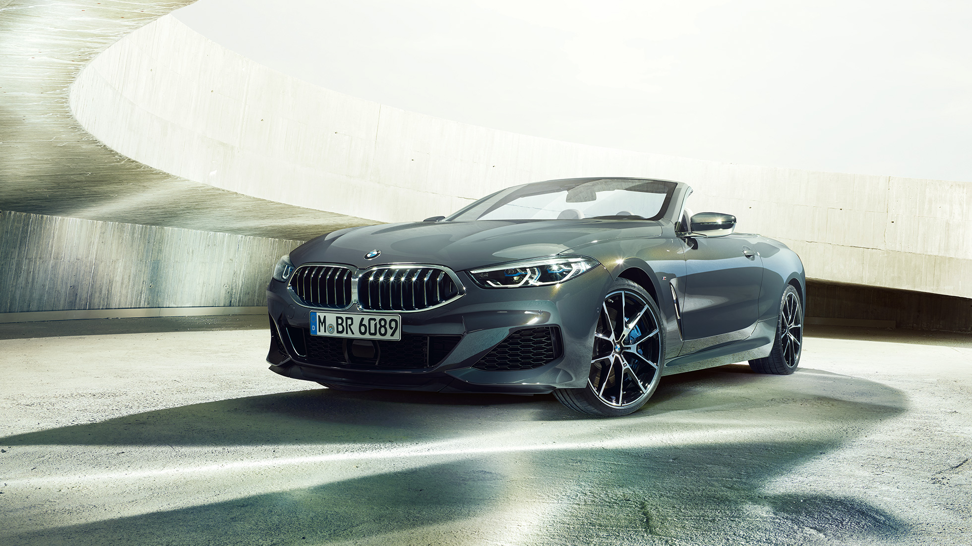 BMW M850i xDrive Cabrio, μπροστινή λήψη τριών τετάρτων. BMW M850i xDrive Cabrio σε γκρι Dravit μεταλλικό με ανοικτή υφασμάτινη οροφή, μπροστινή λήψη τριών τετάρτων, σταθμευμένη μπροστά από τσιμεντένιο κτίριο.
