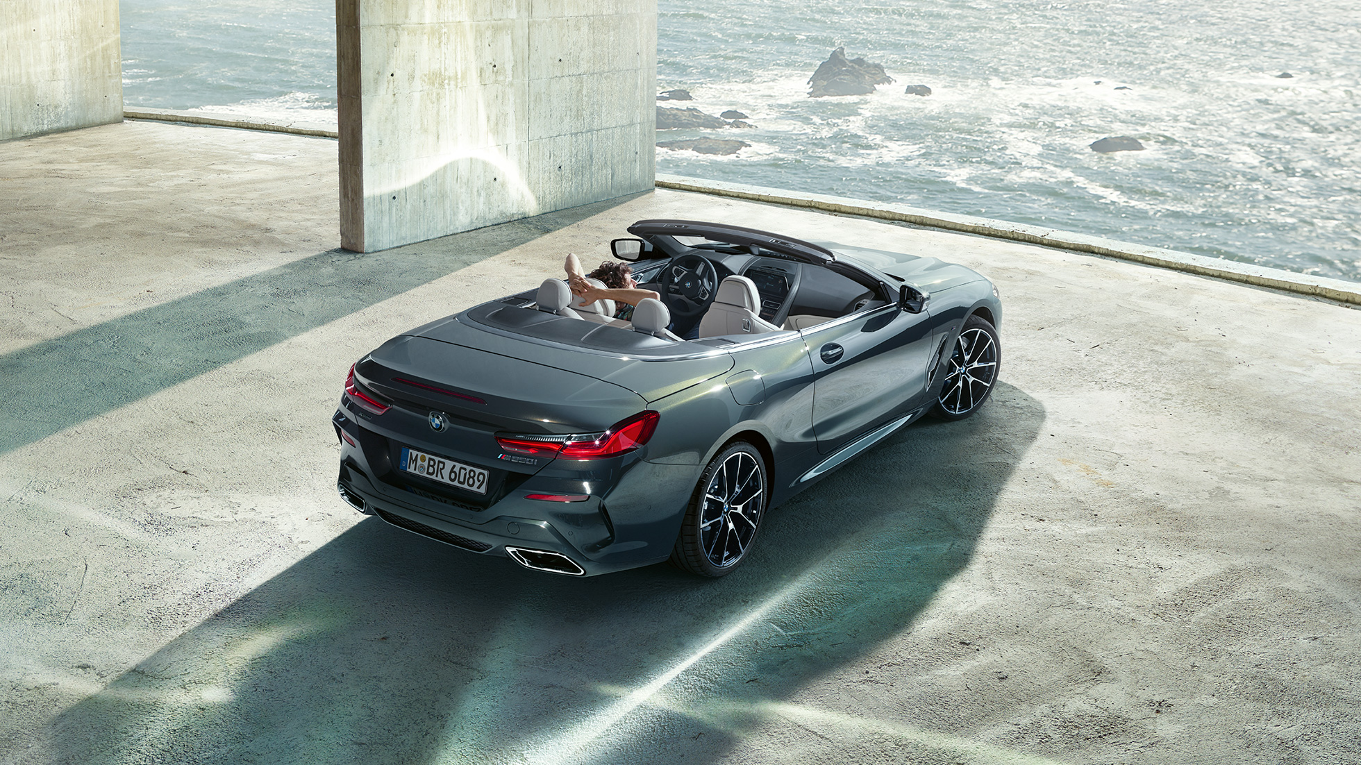 BMW M850i xDrive Cabrio, πανοραμική λήψη. BMW M850i xDrive Cabrio σε γκρι Dravit μεταλλικό με ανοικτή υφασμάτινη οροφή, πανοραμική λήψη, σταθμευμένη σε τσιμεντένιο κτίριο, μπροστά από ωκεανό.