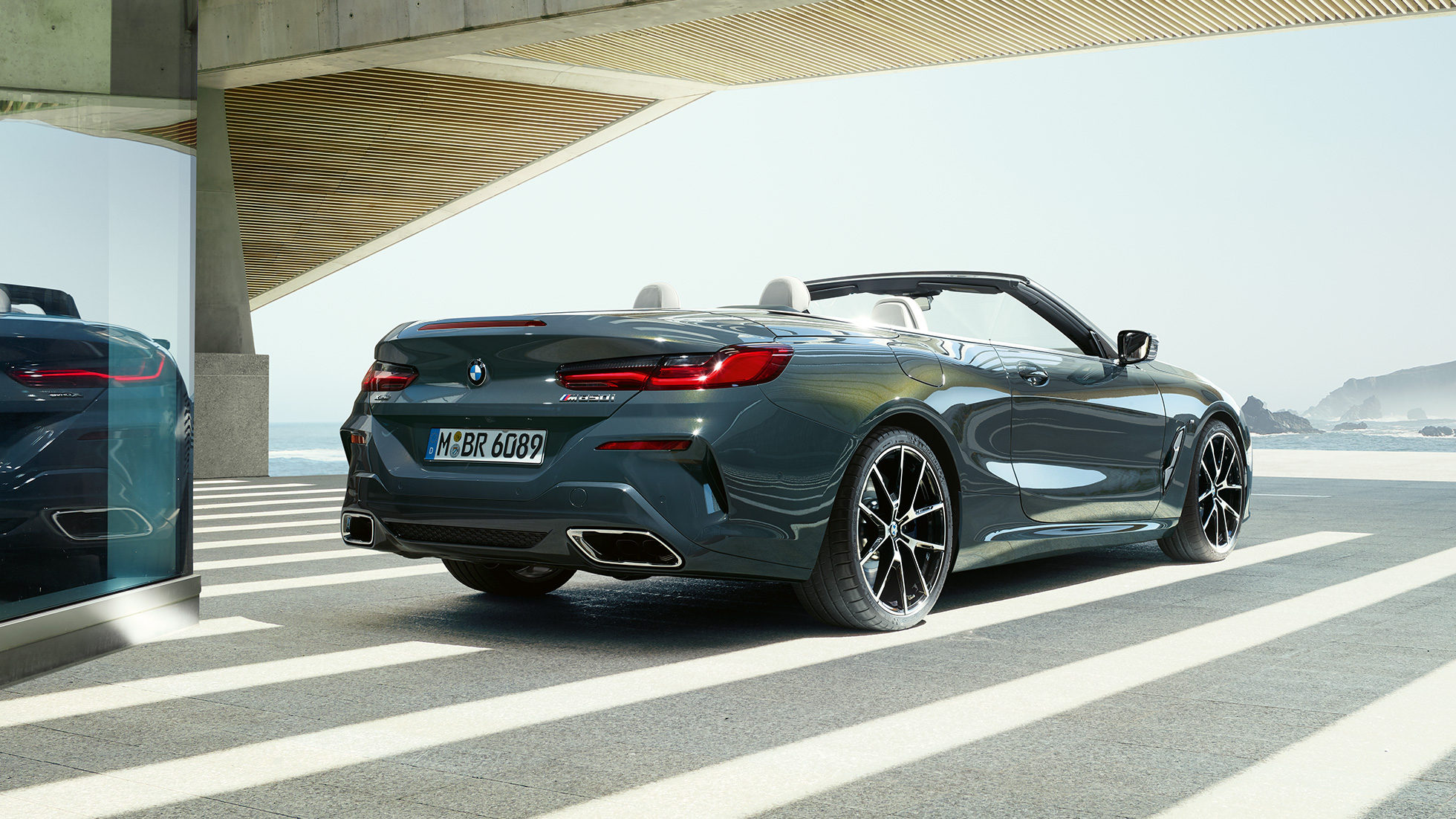 BMW M850i xDrive Cabrio, πίσω λήψη τριών τετάρτων. BMW M850i xDrive Cabrio σε γκρι Dravit μεταλλικό με ανοικτή υφασμάτινη οροφή, πίσω λήψη τριών τετάρτων, σταθμευμένη μπροστά σε ωκεανό.