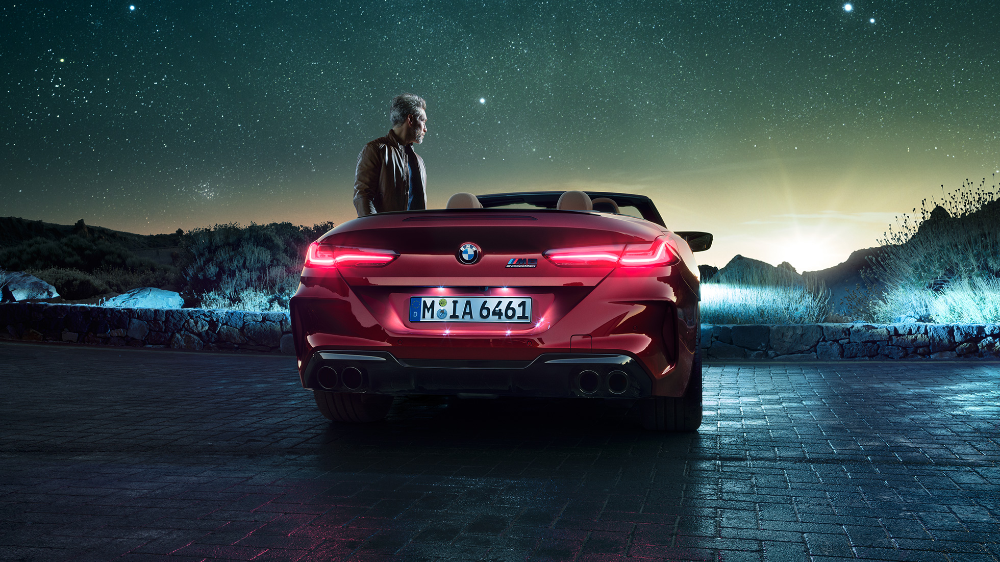 BMW M8 Competition Cabrio, πίσω λήψη. BMW Μ8 Competition Cabrio σε κόκκινο Motegi μεταλλικό με ανοικτή υφασμάτινη οροφή, πίσω λήψη, σταθμευμένη, με άντρα κάτω από έναστρο ουρανό.