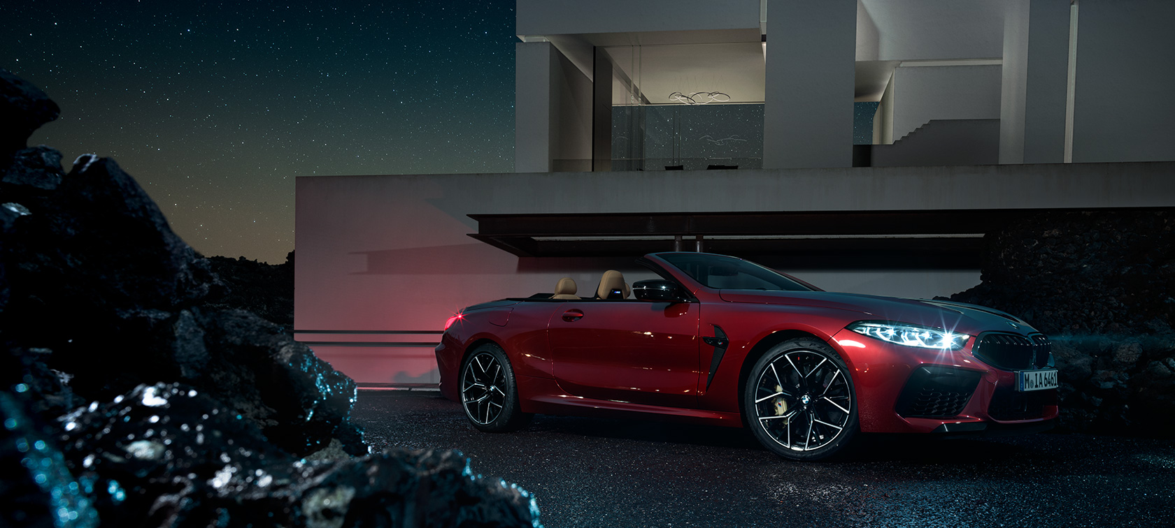 BMW Μ8 Competition Cabrio, πλαϊνή λήψη τριών τετάρτων. BMW Μ8 Competition Cabrio σε κόκκινο Motegi μεταλλικό, πλαϊνή λήψη τριών τετάρτων τη νύχτα, σταθμευμένη μπροστά από μία έπαυλη.
