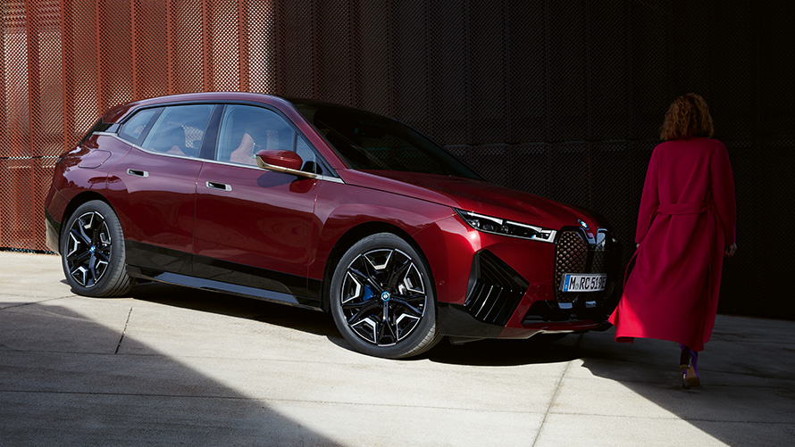 Γυναίκα με κόκκινο παλτό μπροστά από την BMW iX i20 2021 λήψη τρία τέταρτα μπροστά Γυναίκα με κόκκινο παλτό μπροστά από την BMW iX i20 2021 λήψη τρία τέταρτα μπροστά
