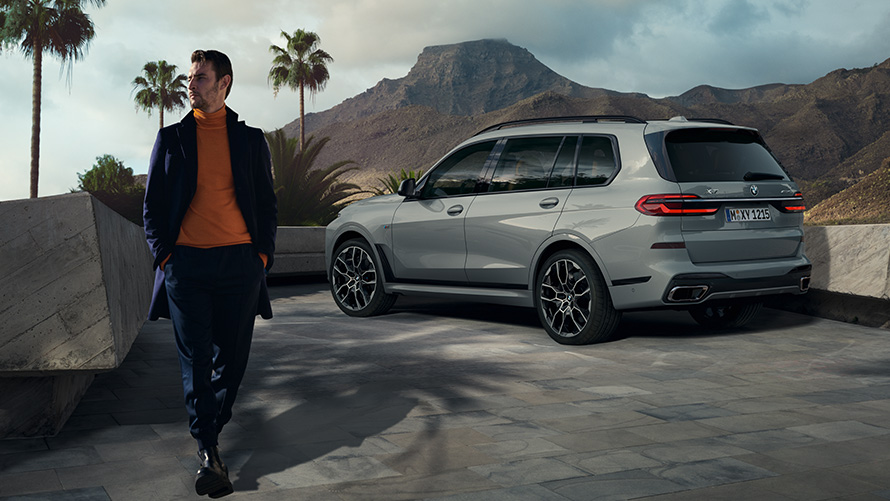Άντρας μπροστά από σταθμευμένη BMW X7 (G07), Απομακρυσμένος Έλεγχος Στάθμευσης Απομακρυσμένος Έλεγχος Στάθμευσης, άντρας μπροστά από σταθμευμένη BMW X7 (G07), πίσω λήψη τριών τετάρτων, κάτω από φοινικιές με ορεινό τοπίο στο φόντο