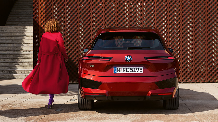 Γυναίκα με κόκκινο παλτό, δίπλα από μία BMW iX (i20), κλείσιμο της πόρτας χώρου αποσκευών Κλείσιμο της πόρτας χώρου αποσκευών, γυναίκα με κόκκινο παλτό δίπλα από μία BMW iX (i20), μπροστά από κτίριο