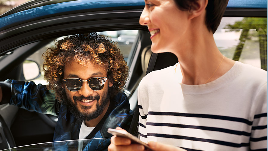 BMW Connected App Άνδρας μέσα στην BMW και γυναίκα έξω από την BMW χρησιμοποιούν ένα smartphone με BMW Connected
