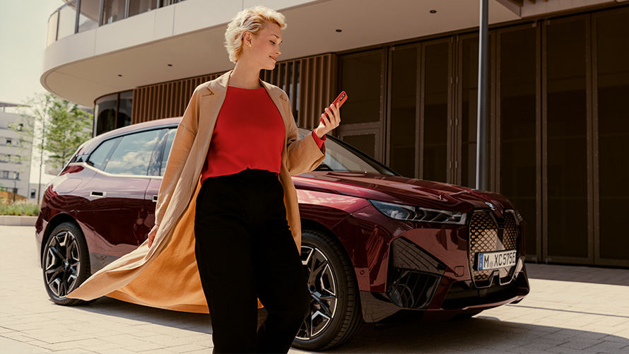My BMW App Εφαρμογή My BMW, BMW iX i20, γυναίκα περπατά μπροστά από ένα αυτοκίνητο BMW κρατώντας ένα smartphone
