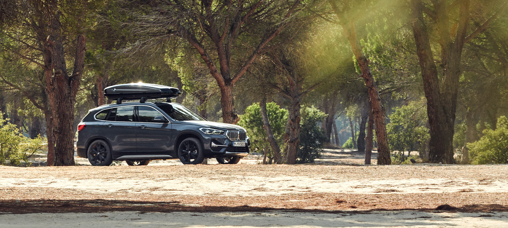 Πλαϊνή λήψη BMW X1 με μπαγκαζιέρα οροφής BMW, προϊόντα καλοκαιρινών προτάσεων BMW Πλαϊνή λήψη BMW X1, σε Mineral Grey με μπαγκαζιέρα οροφής BMW, σταθμευμένη σε δάσος, προϊόντα καλοκαιρινών προτάσεων BMW