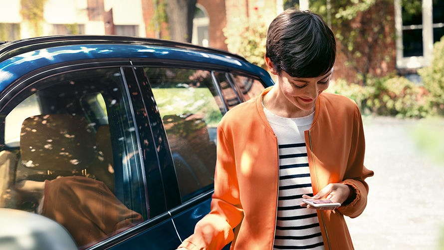 Προϊόντα καλοκαιρινών προσφορών BMW, BMW Teleservices Προϊόντα καλοκαιρινών προτάσεων BMW, BMW Teleservices, γυναίκα στέκεται μπροστά από μια BMW και χρησιμοποιεί το smartphone