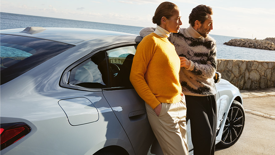 BMW Service Inclusive, διάρκεια και συμβατικές περίοδοι ισχύος για νέα αυτοκίνητα BMW Service Inclusive, άντρας και γυναίκα στηρίζονται στην BMW i4 μπροστά σε ακτή
