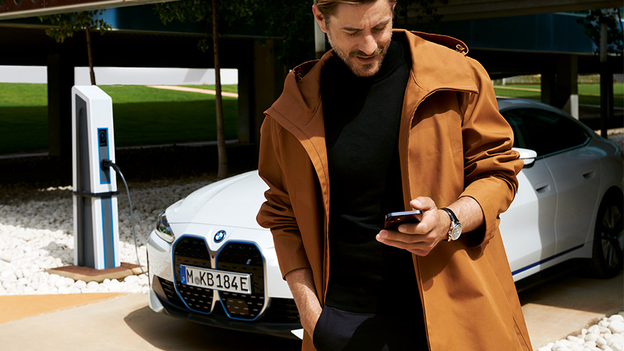 BMW Service Inclusive, διάρκεια και συμβατικές περίοδοι ισχύος για ηλεκτρικά αυτοκίνητα BMW Service Inclusive, άντρας κοιτάζει στο smartphone μπροστά από μία BMW i4 σε σταθμό φόρτισης