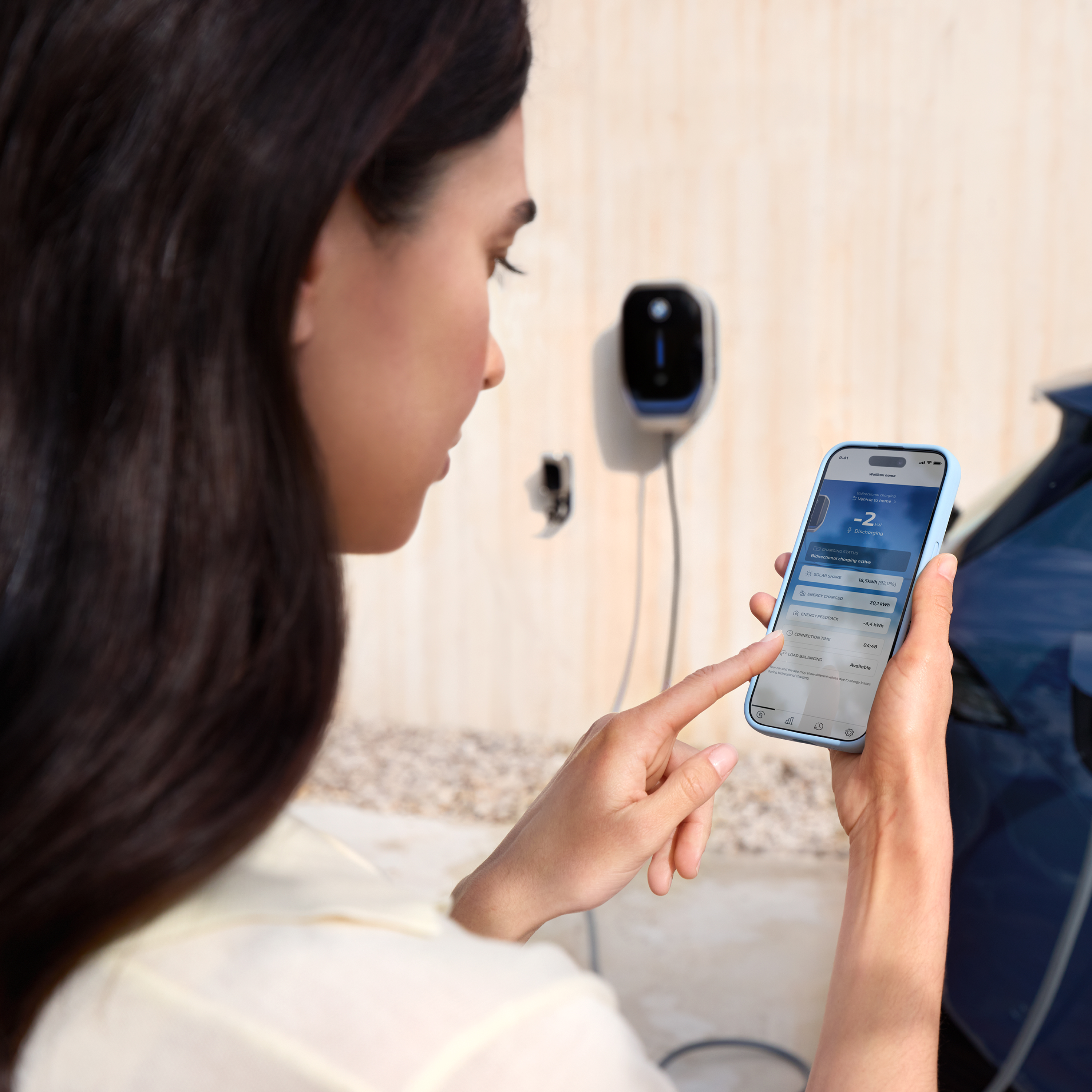 Μια γυναίκα κοιτάζει το smartphone της, ένα Wallbox είναι ορατό στο παρασκήνιο