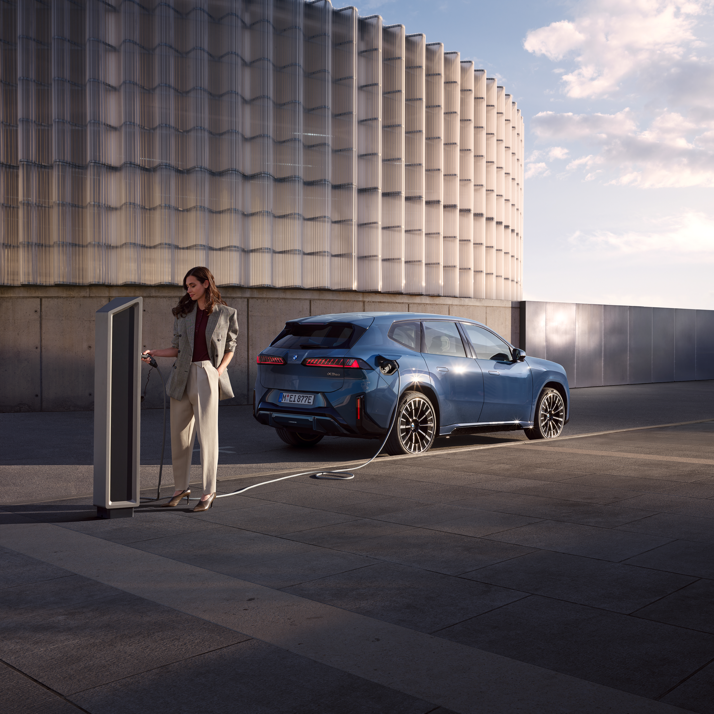 Μια γυναίκα στέκεται δίπλα σε μια BMW iX3, η οποία είναι συνδεδεμένη σε δημόσιο σταθμό φόρτισης